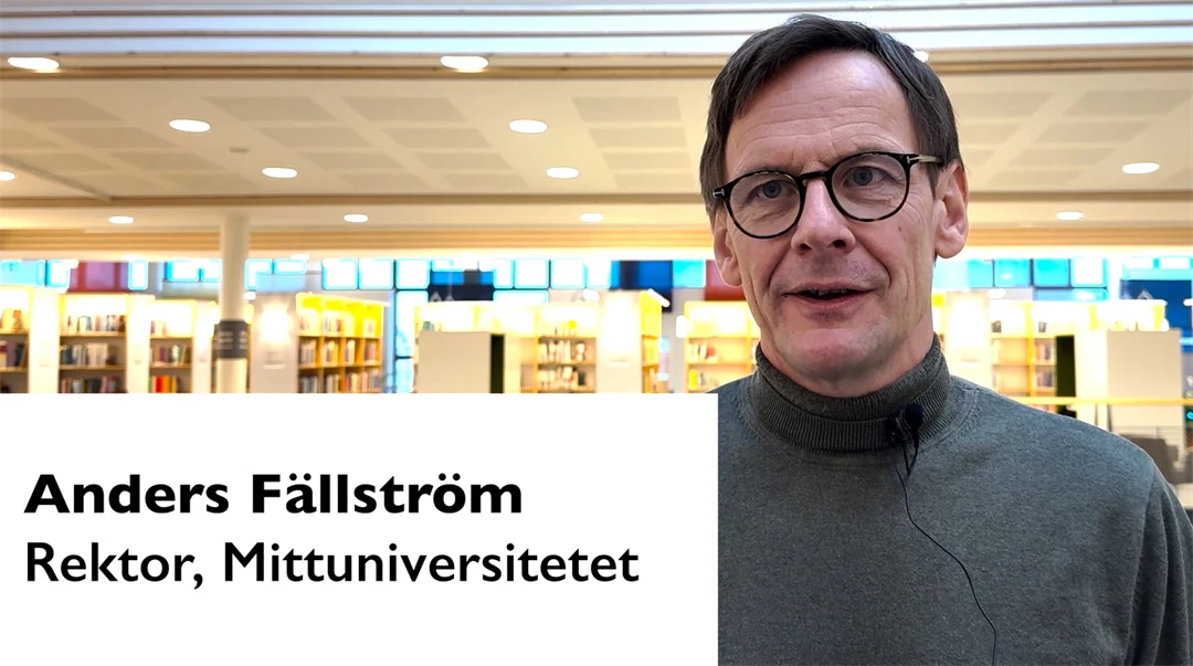 Man med glasögon, mörkt kortklippt hår, klädd i grå polo,  som står i ett bibliotek