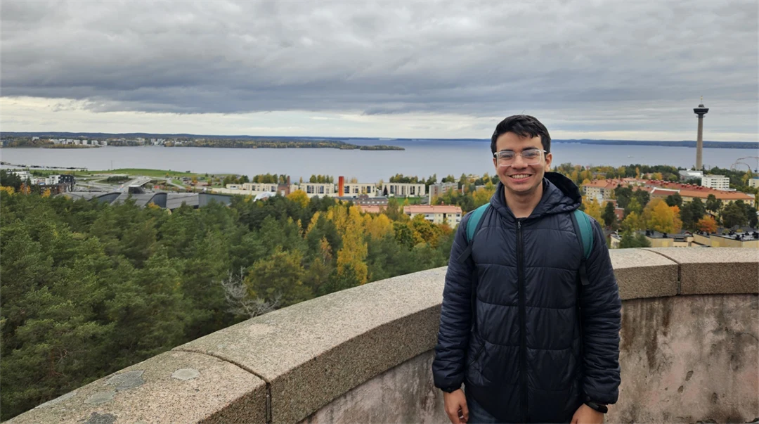 A young man wearing a dark blue jacket and carrying a rucksack is standing by a stone wall high up, looking out over trees, buildings and water. 
