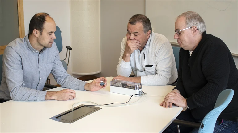 Tre män sitter vid ett bord och diskuterar en prototyp som ligger på bordet.