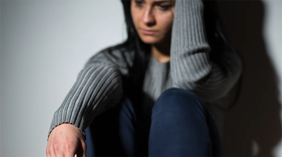 Kvinna klädd i jeans och en grå tröja sitter lutad mot en vägg och ser ledsen ut.