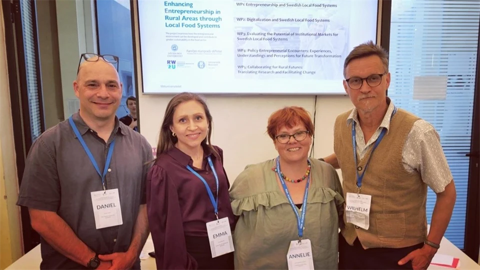 Four people, two men and two women, are standing in a room in front of a screen with a presentation on it. Around their necks are blue lanyards with name tags.