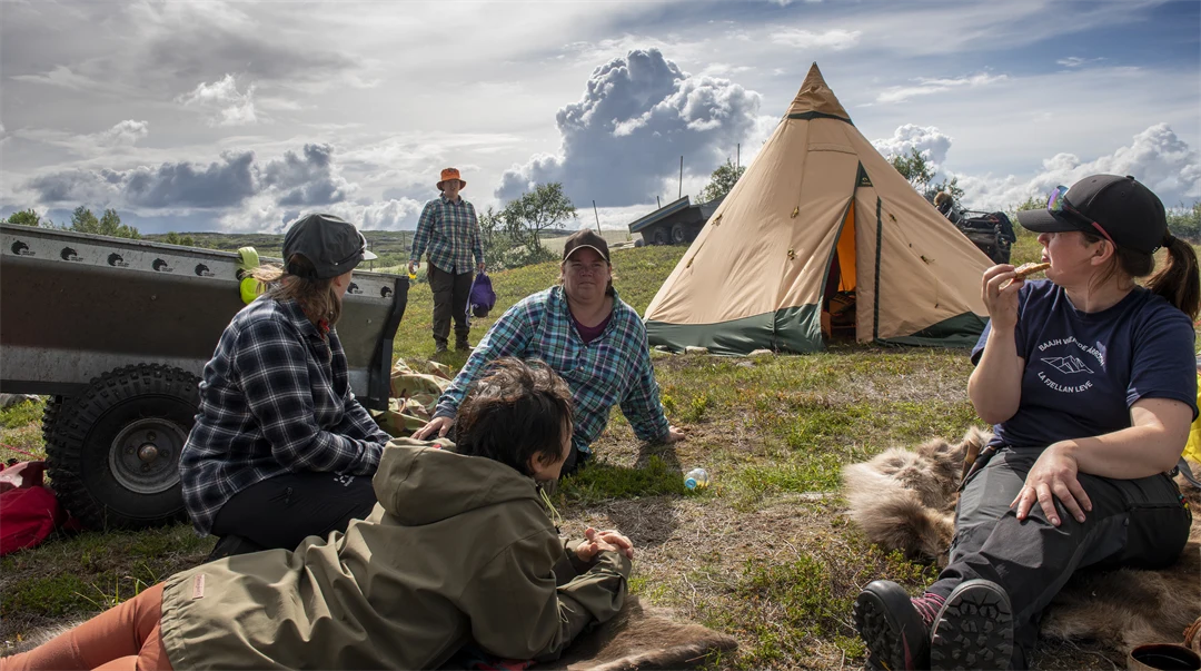 En grupp ungdomar sitter på renskinn på gräset utanför en tältkåta.