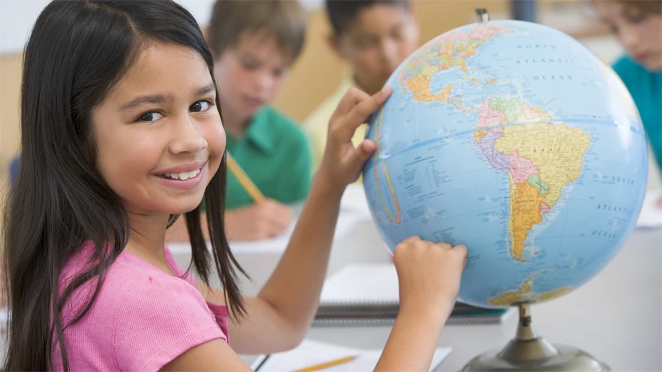 Fyra barn sitter vid ett bord. Tre av dem skriver och en av dem, en flicka med långt mörkt hår och rosa t-shirt pekar på jordglob.
