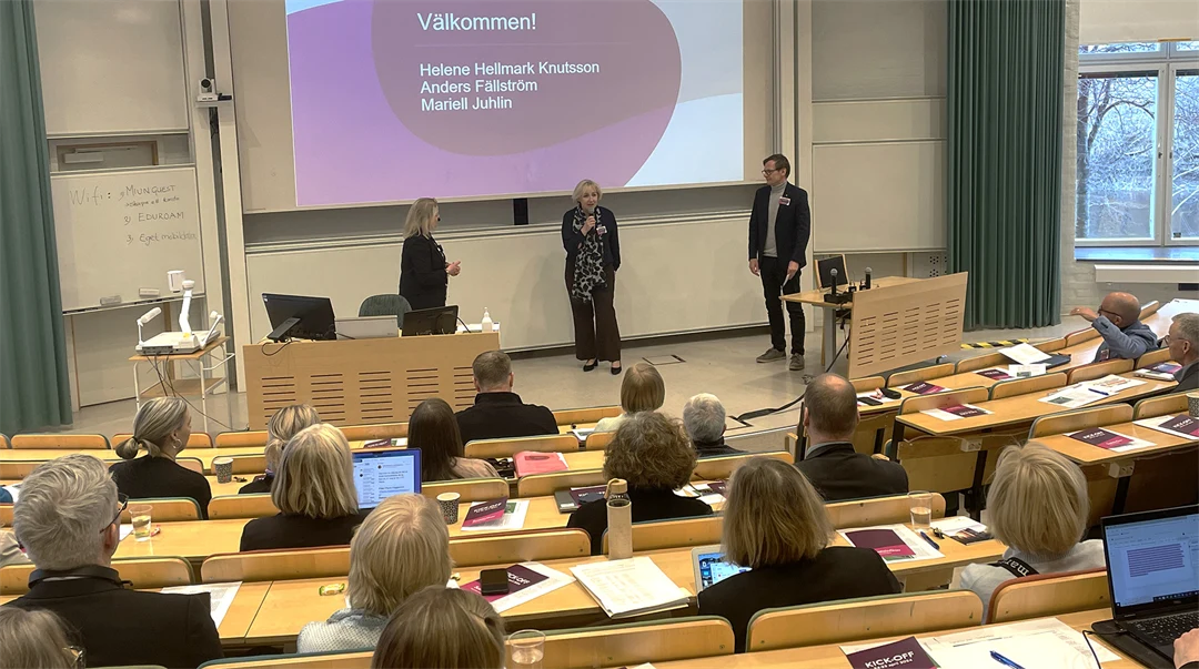 Tre personer står på ett podium framför människor som sitter och lyssnar.