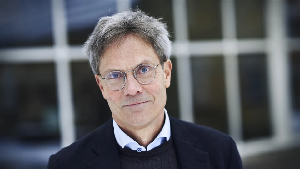 En man i glasögon och mörk kavaj står framför ett glasfönster.