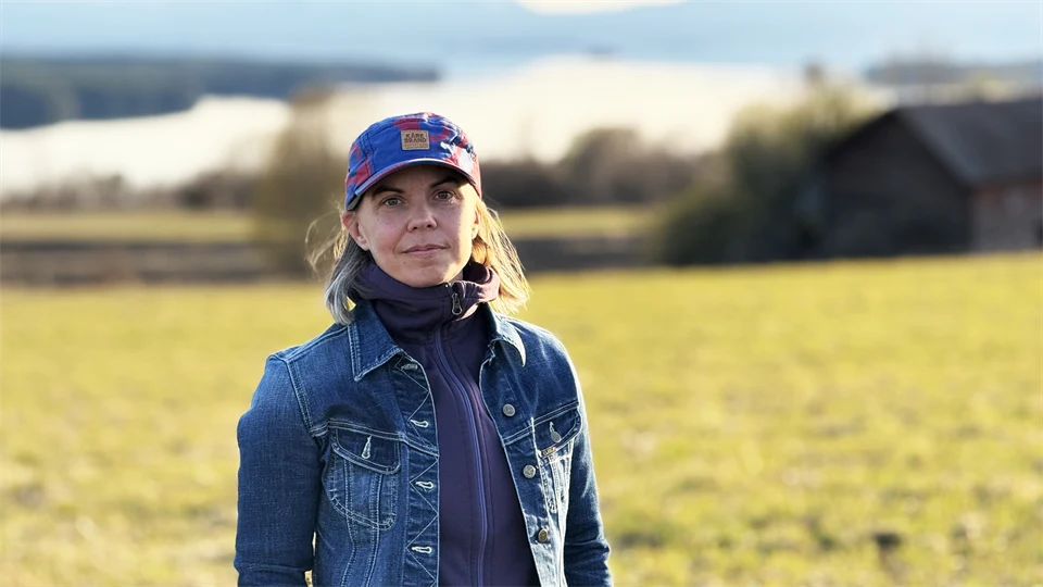 En kvinna i jeansjacka och keps står på en äng. I bakgrunden syns en sjö och fjäll.
