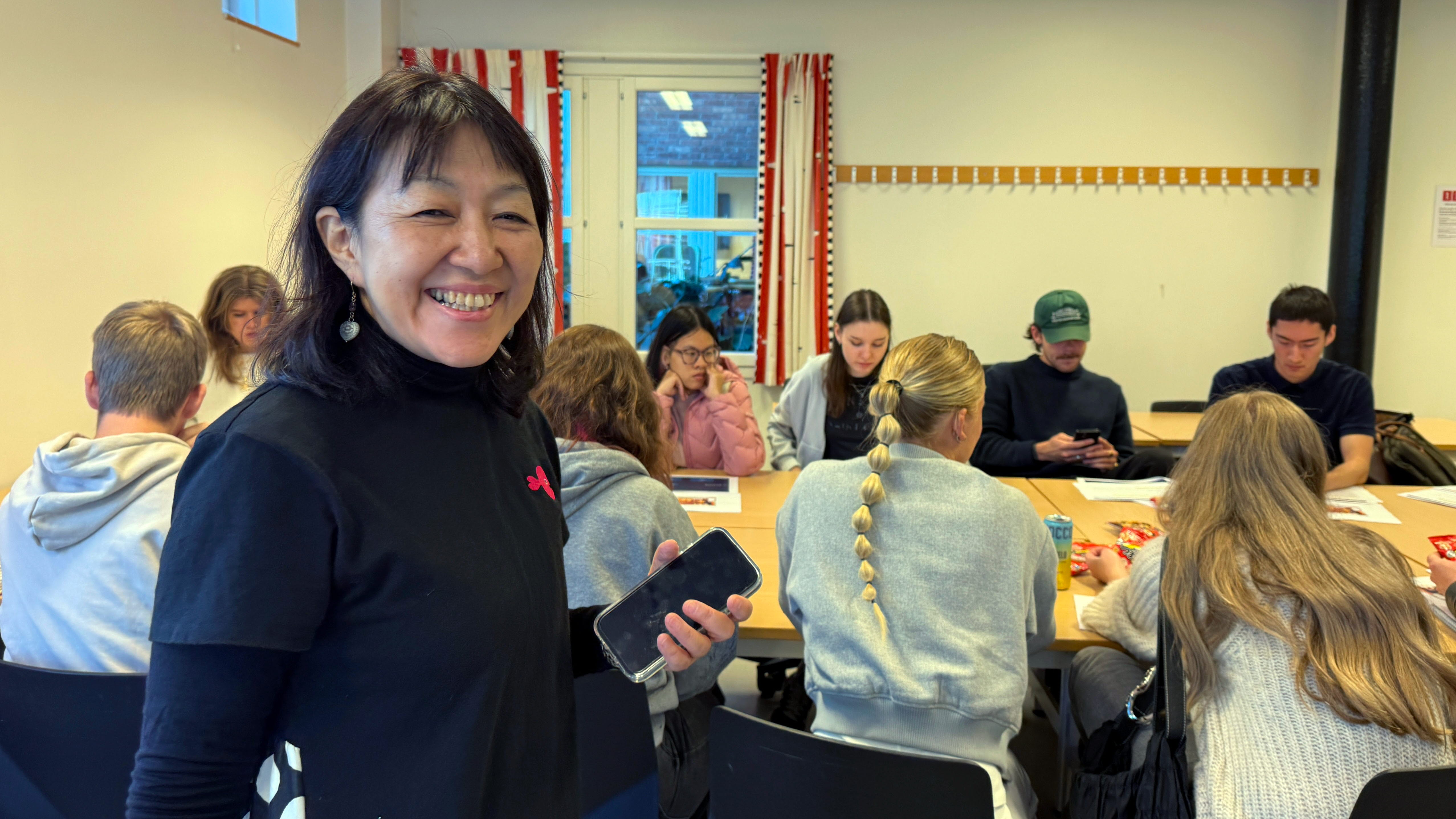 En kvinna står i ett klassrum, studenter sitter i bakgrunden