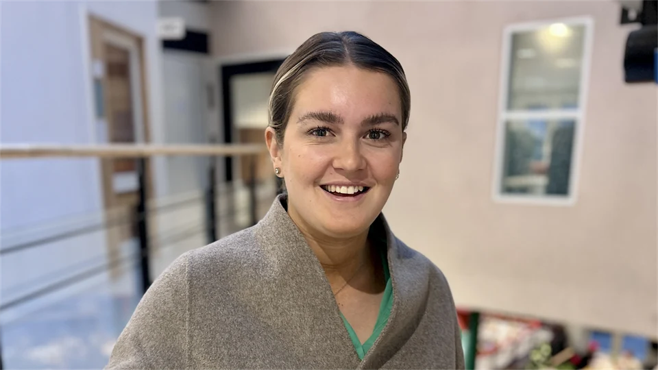 En mörkblond kvinna klädd i beige kofta står i trappa med beige vägg bakom sig.