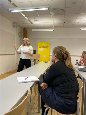 En kvinna i vit jacka och svarta byxor står längst fram i ett klassrum och pekar. Några personer sitter på stolar och lyssnar med block och pennor framför sig.
