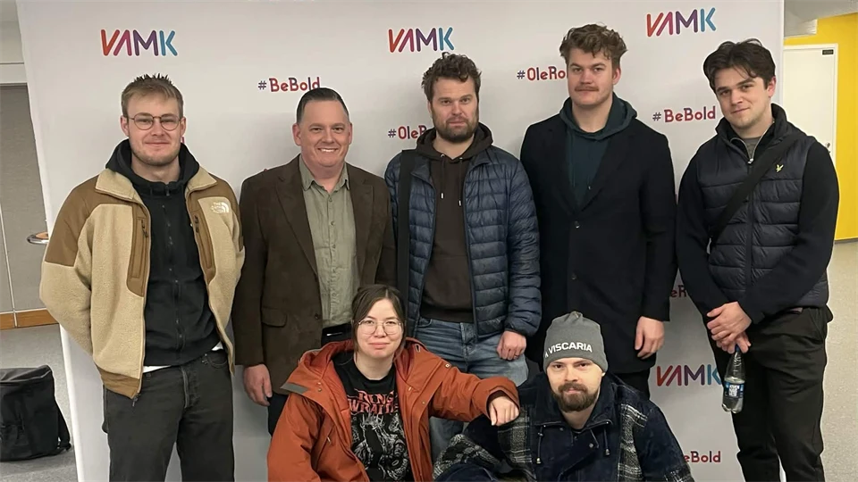 Seven people pose for a group photo in front of a white backdrop with VAMK logos. Five are standing and two is kneeling in front. They are wearing casual outerwear in a indoor setting.