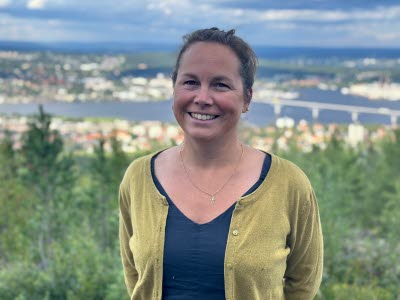 En kvinna med uppsatt brunt hår klädd i grön kofta och blå tröja står framför ett naturlandskap med utsikt över skog och hav. 