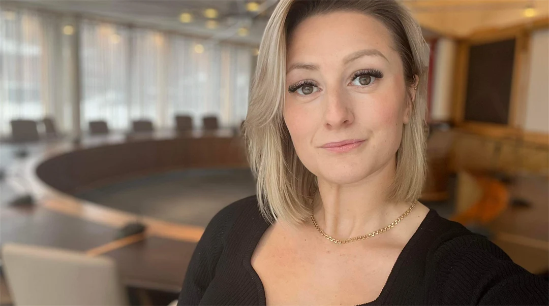 Kvinna med ljust och kort hår tittar in i kameran, kvinnan bär en svart stickad tröja och står framför ett stort mötesbord.