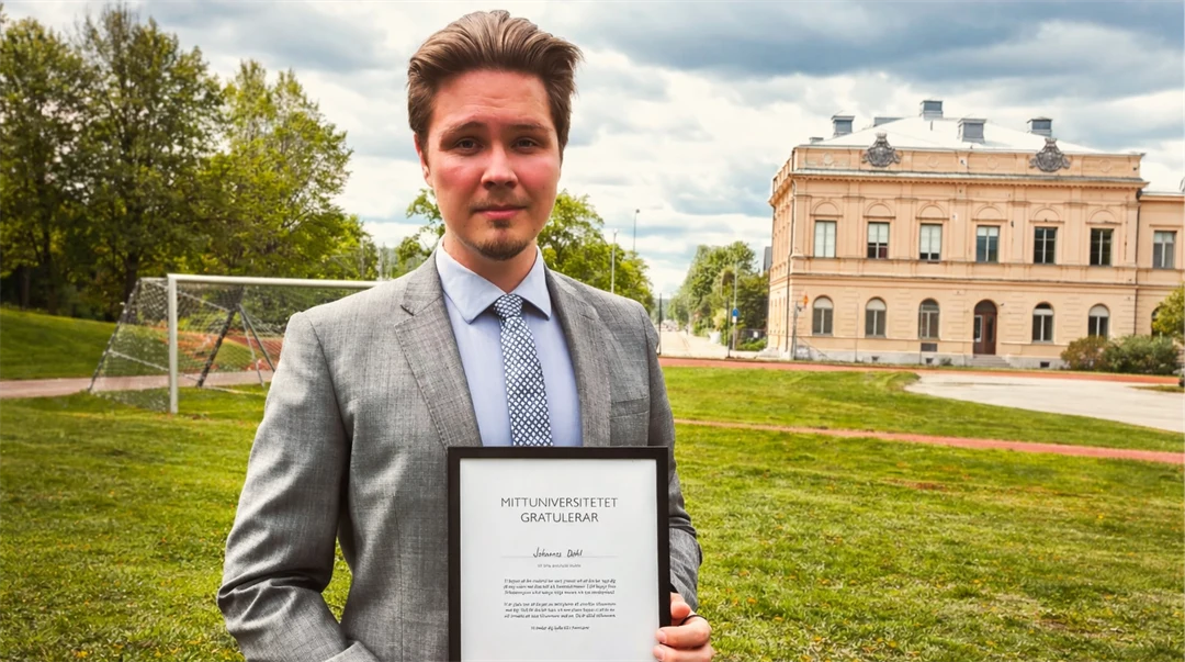 En man i grå kostym håller i ett diplom. I bakgrunden finns en gräsmatta och en byggnad.