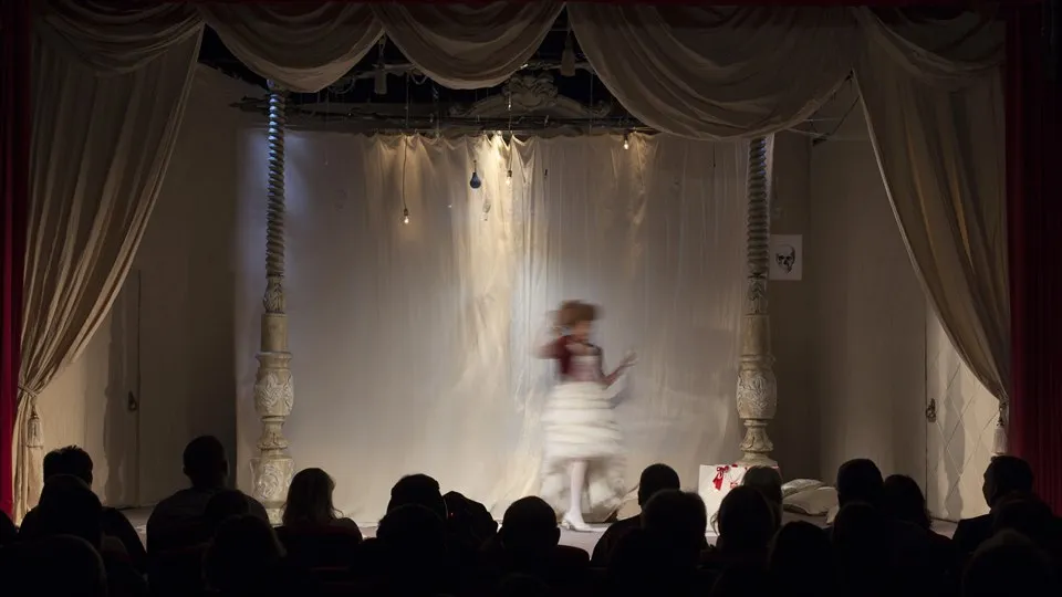 teaterscen med publik i förgrunden. På scenen står en kvinna.