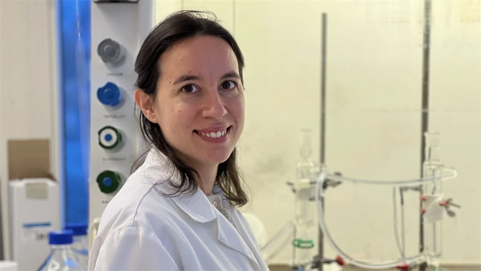 A woman looks into the camera and smiles. In the background is a panel with knobs and laboratory equipment.