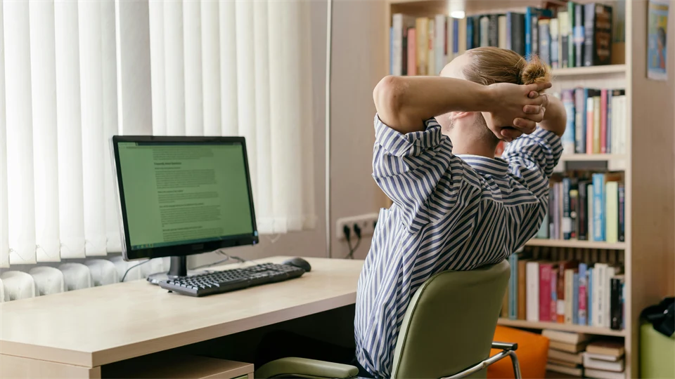 Person lutar sig lite bakåt med händerna bakom huvudet vid ett skrivbord med en datorskärm