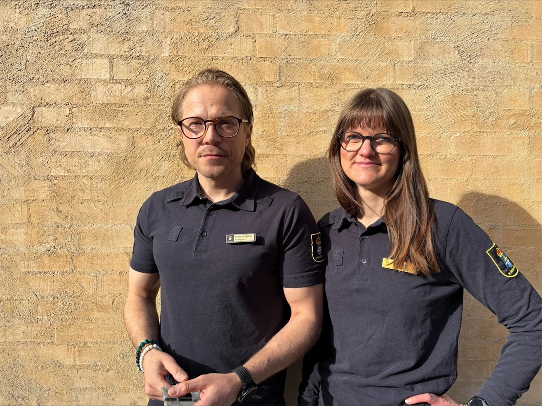 Två personer står framför en beige husfasad iklädda uniformer från Kriminalvården