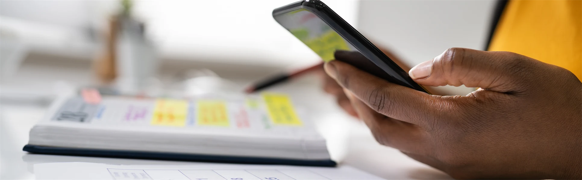 En hand håller i en mobiltelefon och en kalender ligger på ett bord.