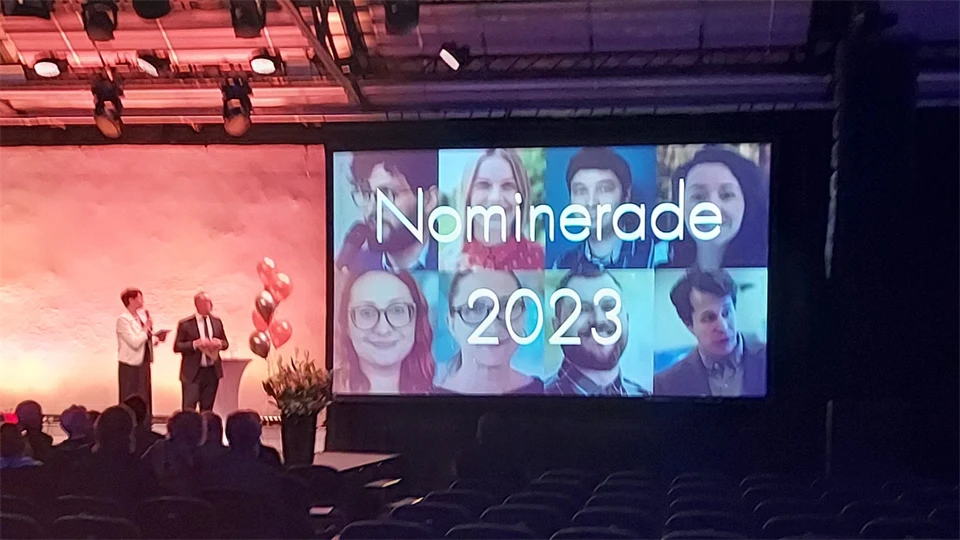 Två personer på en upplyst scen och en stor skärm som visar texten "Norminerade 2023" och åtta ansikten.