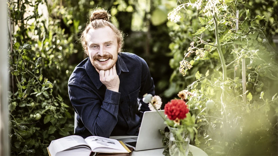 Manlig student sitter i  växthus och pluggar med laptop och böcker. 