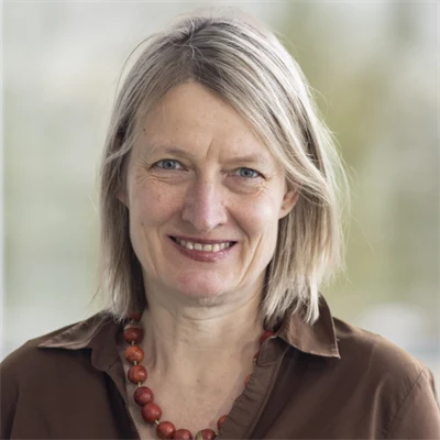 En kvinna i axellångt, blont hår, halsband med röda, stora, runda pärlor och brun skjorta. 