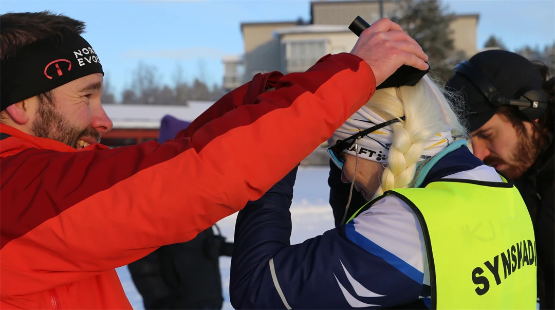 Man i röd jacka hjälper synskadad, kvinnlig skidåkare att bära skallbenshörlurar 