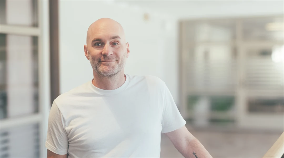En man i en vit t-shirt står i en ljus lokal. Han ler och tittar in i kameran.,