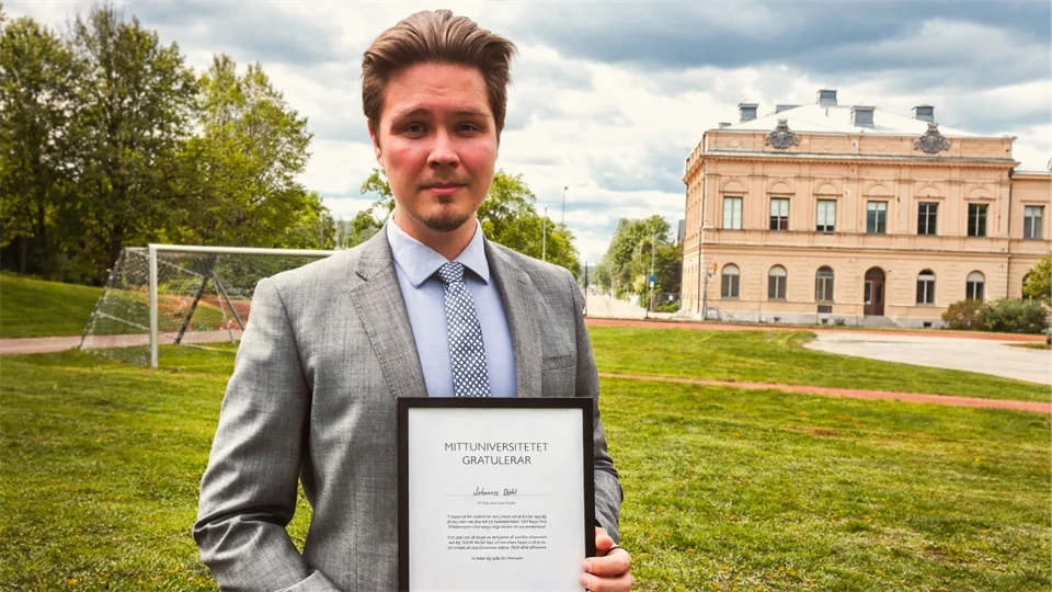 En man i grå kostym håller i ett diplom. I bakgrunden finns en gräsmatta och en byggnad.