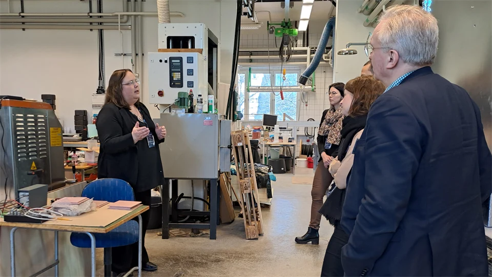 En grupp personer i en verkstad lyssnar på en kvinna som talar.