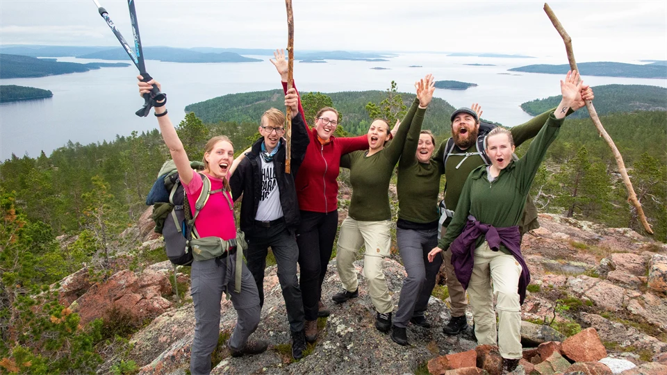 A group of people celebrate having reached the top, in the background a view of forest and water.