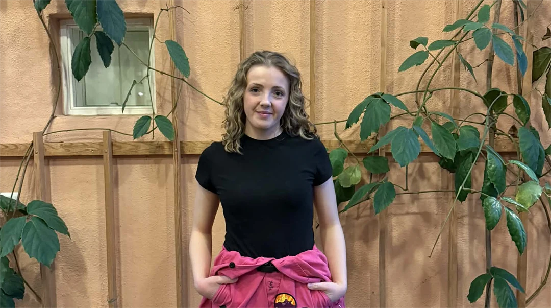 Ung kvinna med svart kortärmad tröja och ceriserosa overall knuten runt midjan står framför en beige vägg med gröna växter som slingrar sig. 