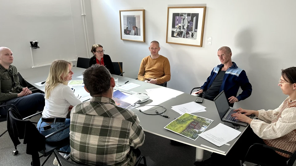 En grupp människor sitter runt ett konferensbord i ett mötesrum. På bordet finns papper, en bärbar dator och en telefon. Två tavlor hänger på väggen bakom dem.