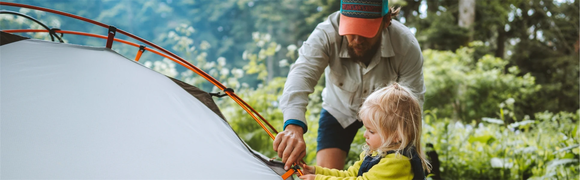 Young man and his daughter, who is about three years old, are setting up a tent in the forest. They are camping and are wearing sporty leisure wear.