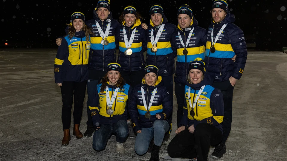 Svenska herr- och damlandslaget i skidskytte står med sina medaljer kring halsen i mörkret på Åre-Östersund Airport.