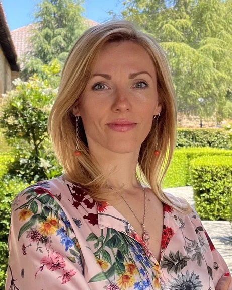 Kvinna med blommig blus står i trädgård.