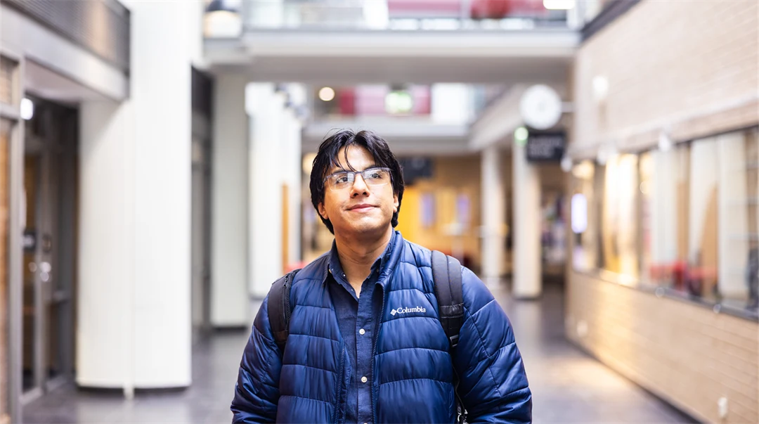 A man with black hair and glasses, wearing a blue jacket and carrying a black backpack, stands in a bright, airy indoor setting and looks past the camera. 