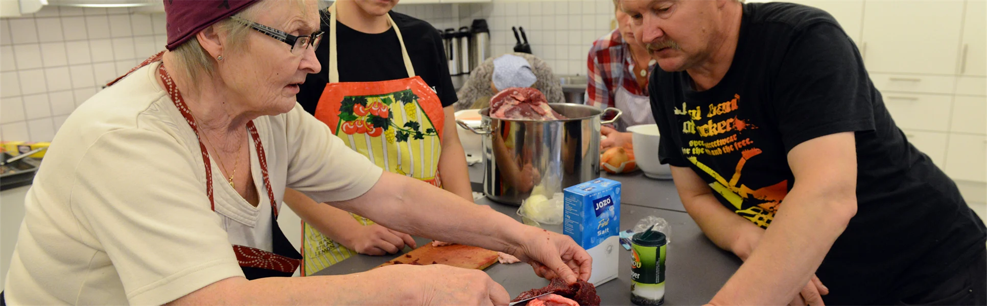 En äldre kvinna visar en ung kvinna och en man hur man tillagar renkött.