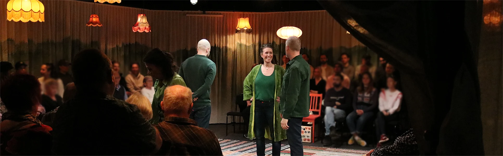 En teaterscen med tre personer i gröna tröjor på och publik runtomkring. 