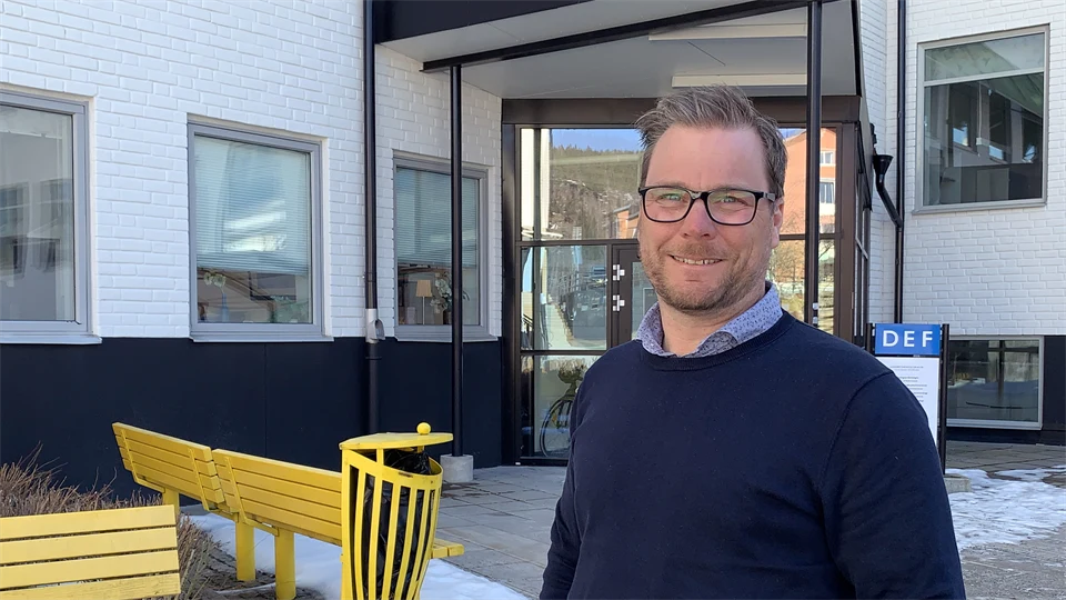 En man i glasögon och marinblå tröja som står utanför ett vitt hus med gula bänkar i bakgrunden