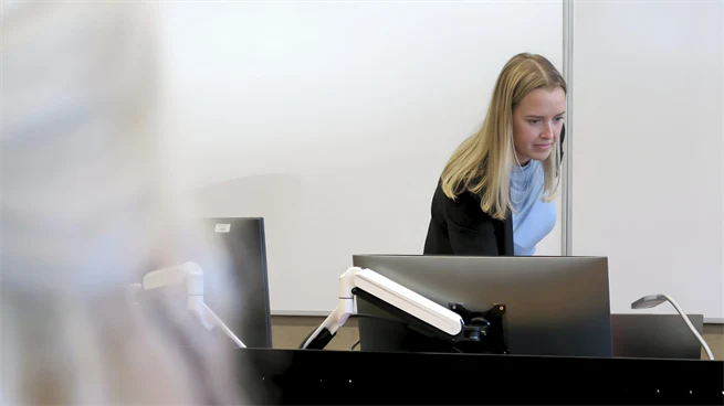 Ung kvinna vid katern i en lektionssal på campus i Östersund. Hon har ljust hår och är klädd i svart kavaj. 