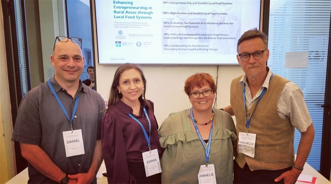 Four people, two men and two women, are standing in a room in front of a screen with a presentation on it. Around their necks are blue lanyards with name tags.