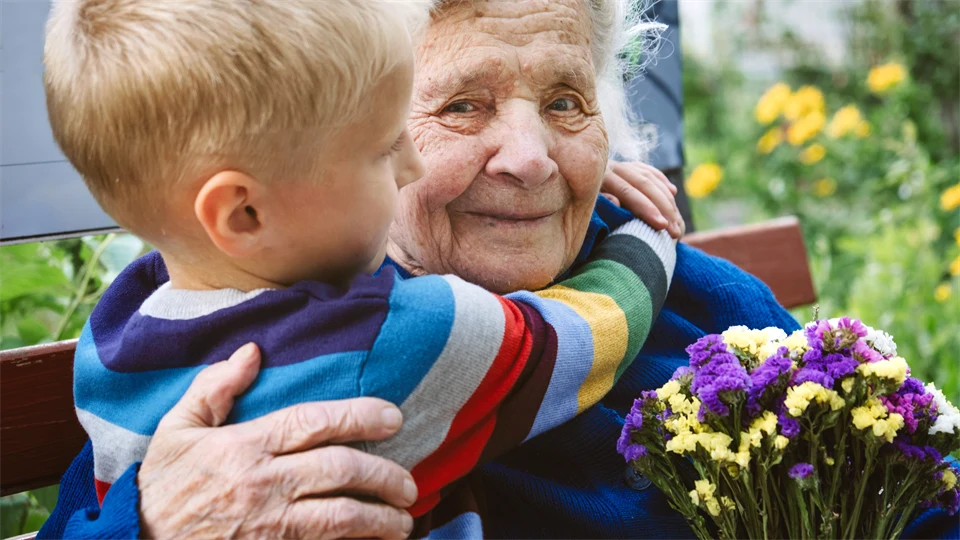 En liten pojke kramar en gammal kvinna, som sitter på en parkbänk med en bukett blommor i handen