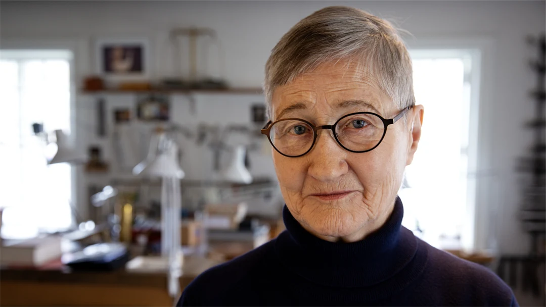 En kvinna med glasögon och kort hår tittar in i kameran. I bakgrunden ser man arbetsbänkar och hyllor.