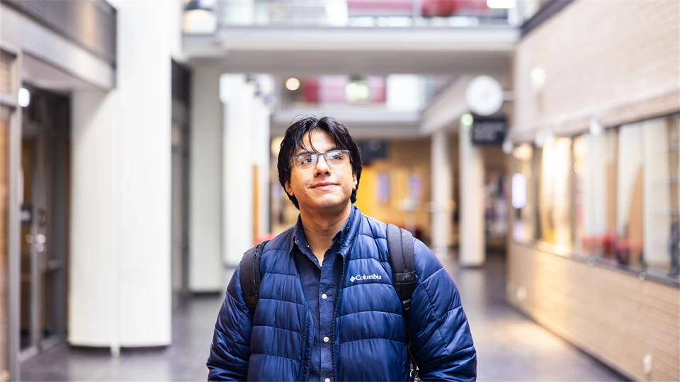 A man with black hair and glasses, wearing a blue jacket and carrying a black backpack, stands in a bright, airy indoor setting and looks past the camera. 