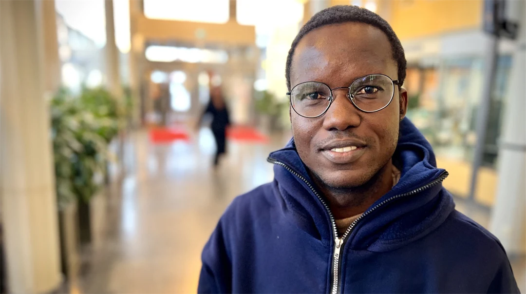 A man is standing in an hall. He is wearing a blue hoodie.